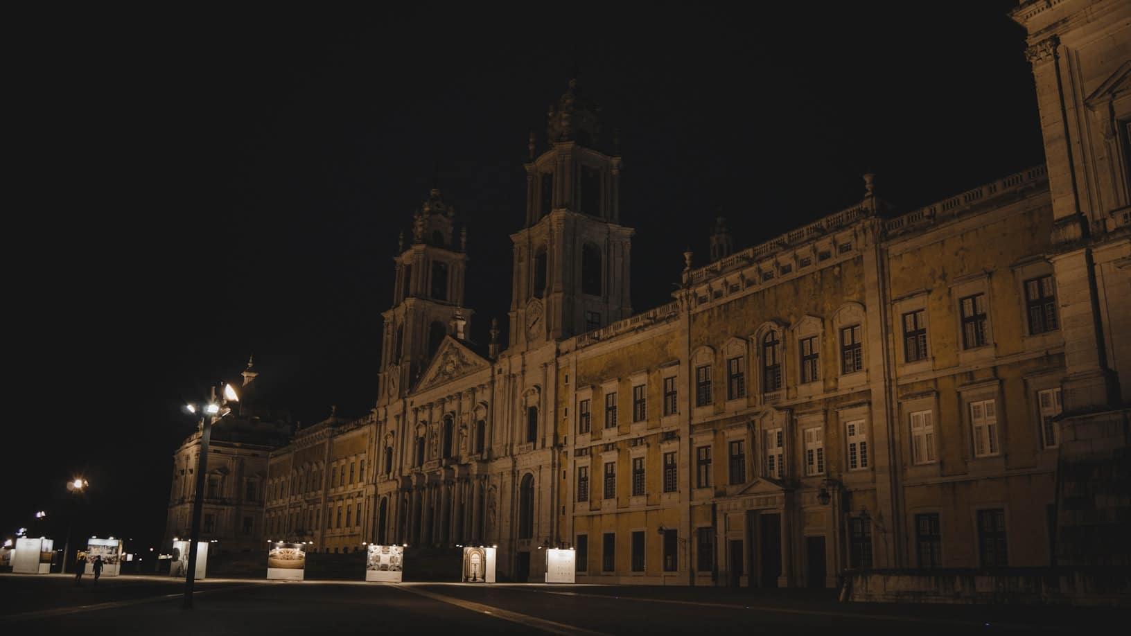 Palácio Nacional de Mafra, grande edifício histórico com duas torres proeminentes, iluminado por candeeiros à noite.