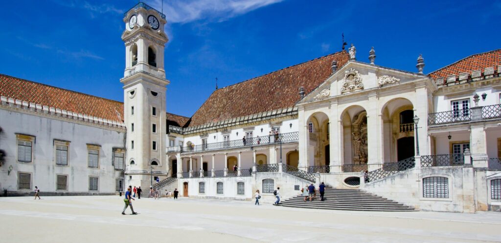 Construção histórica em Coimbra com um pátio à frente, onde pessoas caminham, e uma torre do relógio junto do edifício.