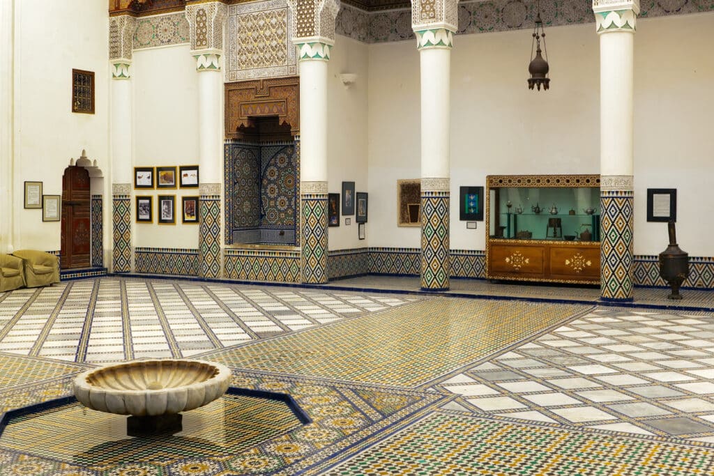 Uma sala com padrões complexos de azulejos, colunas brancas, obras de arte emolduradas nas paredes, uma pequena fonte e uma vitrina, no museu de Marraquexe.