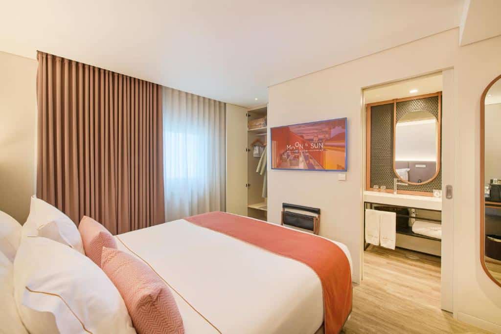 Quarto moderno do Hotel Moon & Sun Braga com cama bem feita, TV de parede, janela com cortinas e zona de casa de banho visível.