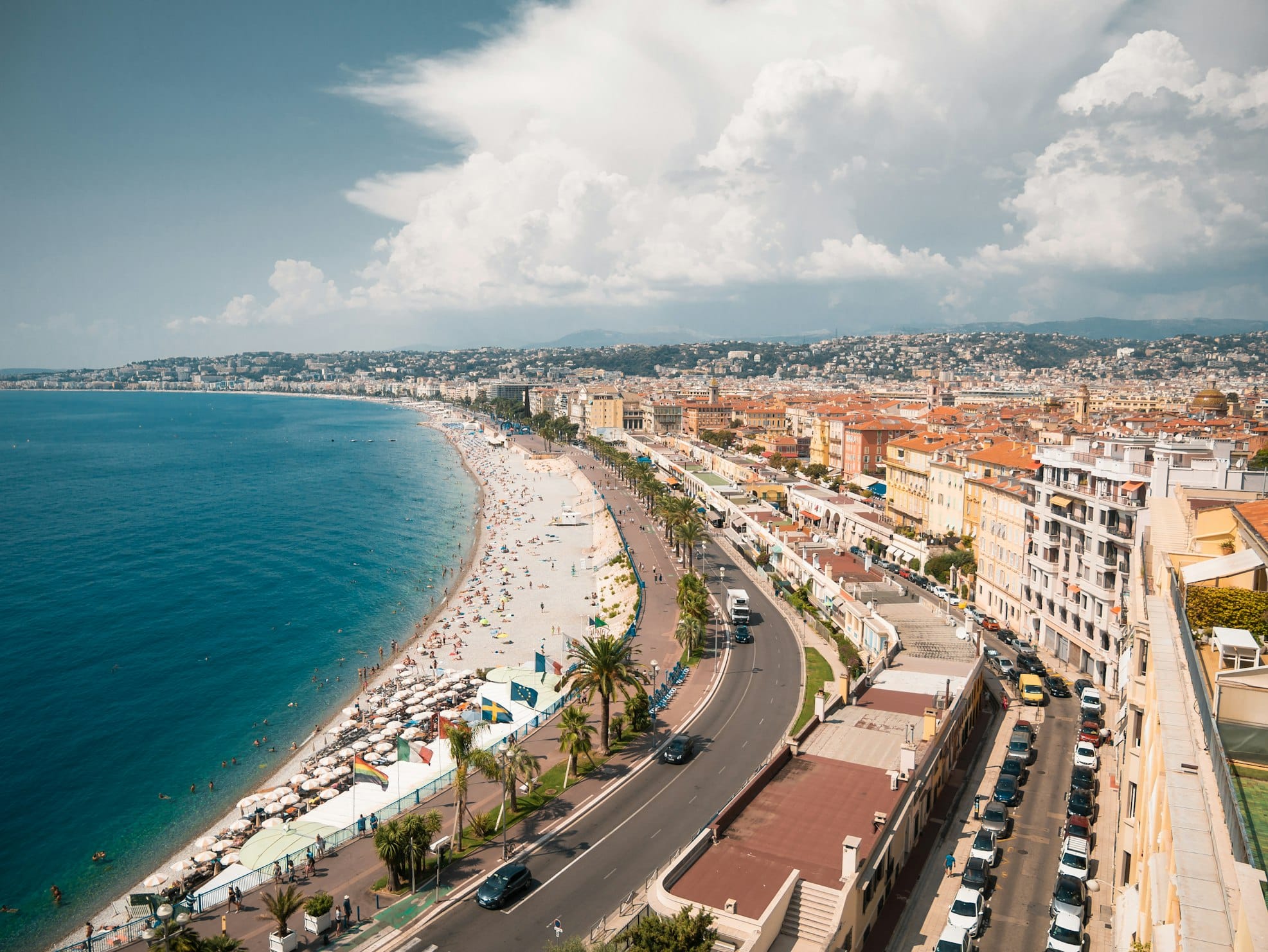 Paisagem costeira de Nice, com praia longa e curva, mar azul e uma avenida ladeada por edifícios e palmeiras.