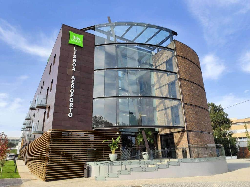 Edifício do Ibis Styles Lisboa Aeroporto com fachada de vidro curvo e exterior castanho.