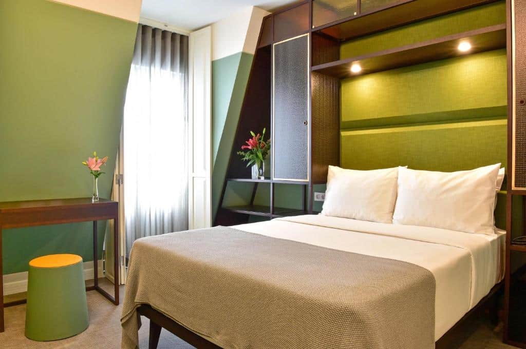 Cama de casal bem arrumada num quarto moderno de tons verdes, com secretária, banqueta e flores junto à janela, no My Story Hotel Augusta.