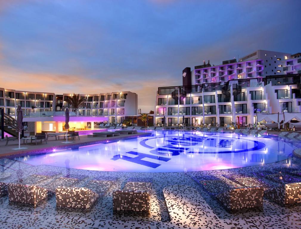 Hard Rock Hotel Ibiza ao anoitecer, com uma grande piscina, iluminação roxa, edifícios de vários andares e espreguiçadeiras junto à piscina.