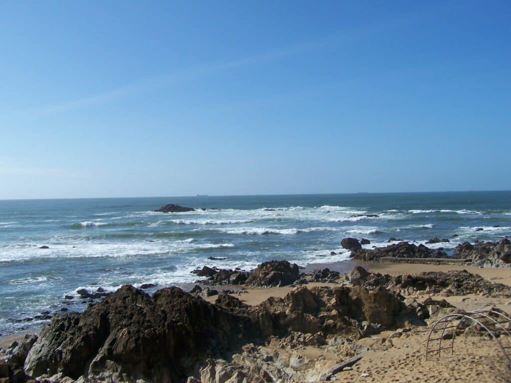 Praia de Matosinhos durante o dia, com areal e rochas à frente e o mar ao fundo.