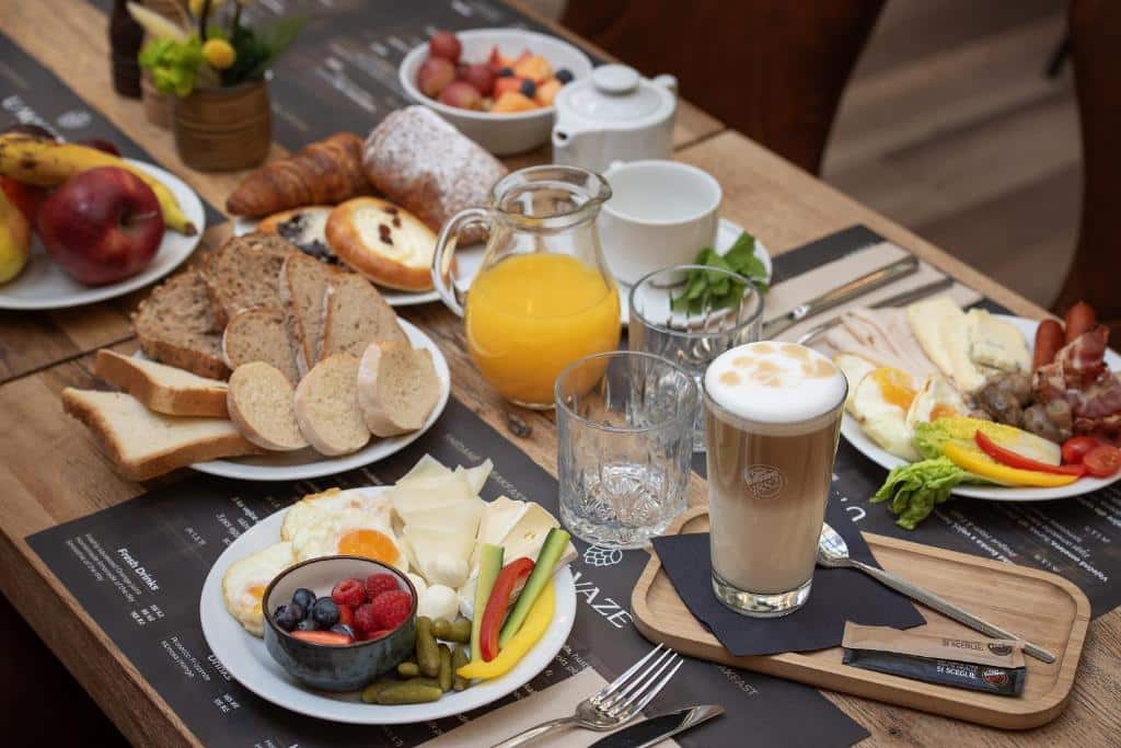 Pequeno-almoço no Little King Hotel U Malvaze, com pão, fruta, queijo, enchidos, ovos, sumo, café e bolos numa mesa de madeira.