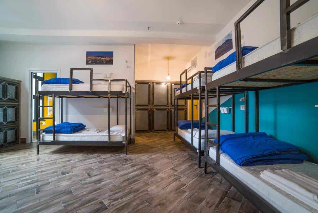 Dormitório de albergue com beliches de metal pretos e lençóis azuis, pavimento de madeira clara e decoração minimalista, no Ostello Bello Napoli.