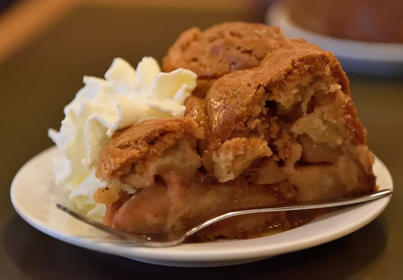 Prato branco com uma fatia de tarte de maçã e natas, com um pequeno garfo ao lado