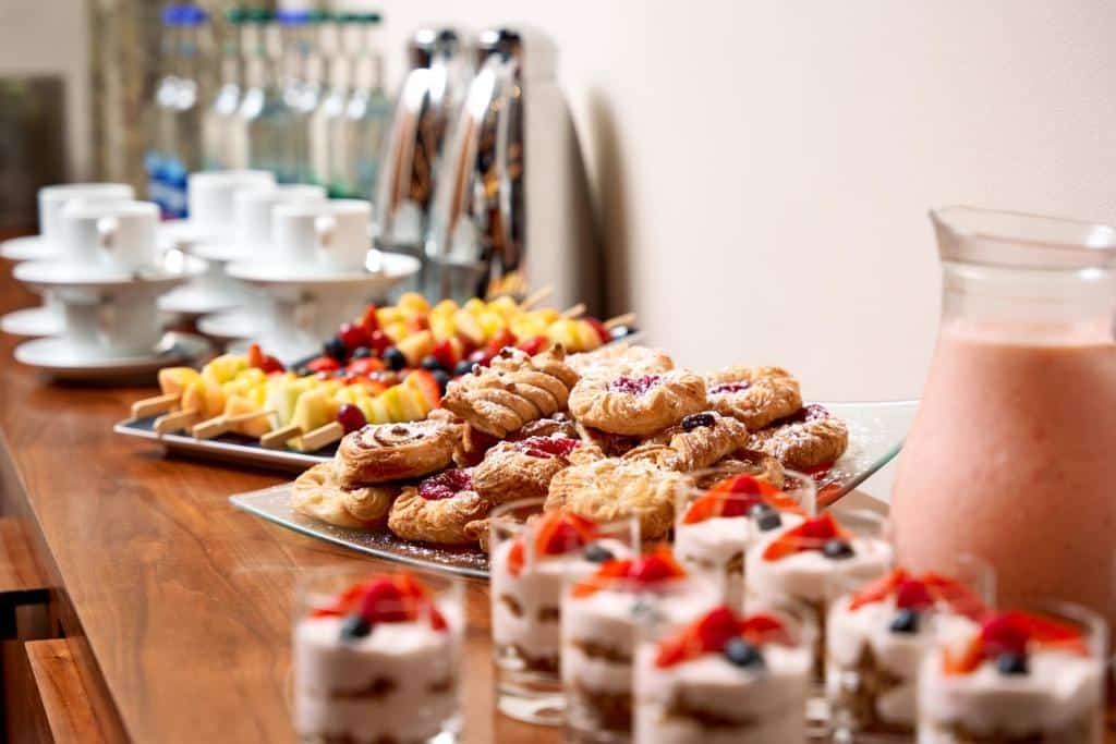 Snacks e fruta no pequeno-almoço do Herbert Park Hotel and Park Residence, incluindo bolachas, tartes e copos com bebida cremosa cor-de-rosa, num ambiente de cozinha acolhedor com prateleiras de vidro.