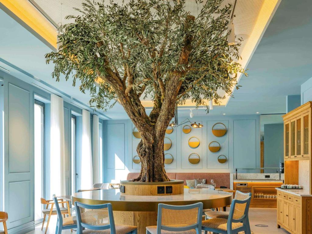 Sala de refeições do Mercure Porto Centro Aliados com mesas e cadeiras e uma árvore ao centro.