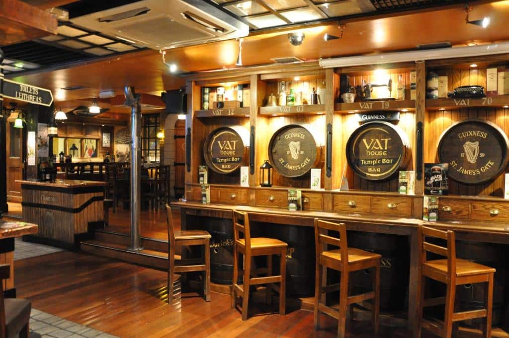 Interior rústico e acolhedor de um bar irlandês tradicional no Blooms Hotel. Mesas de madeira, cadeiras e prateleiras decoradas com barris e garrafas de Guinness, com atmosfera típica de pub e iluminação quente.