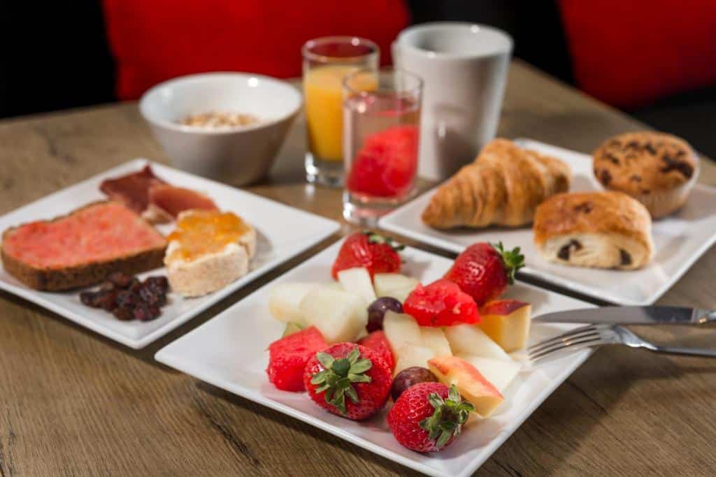 Mesa de pequeno-almoço no Hotel ibis Braga, com fruta, doces, sumo, café, cereais e pães em pratos brancos.