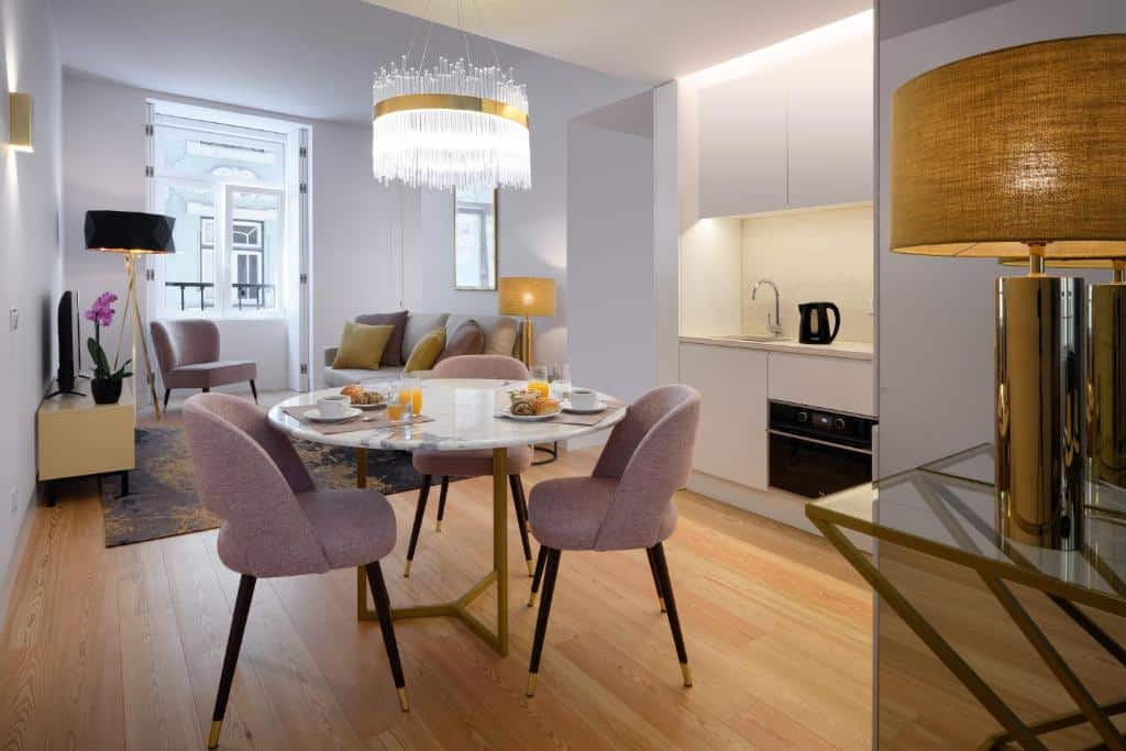 Cozinha em conjunto com uma pequena sala de estar numa das unidades do Ouro Grand by Level Residences, com uma mesa redonda com três cadeiras estofadas, uma janela com televisão e um sofá perto dela e, no canto direito, uma pequena cozinha e um candeeiro de teto