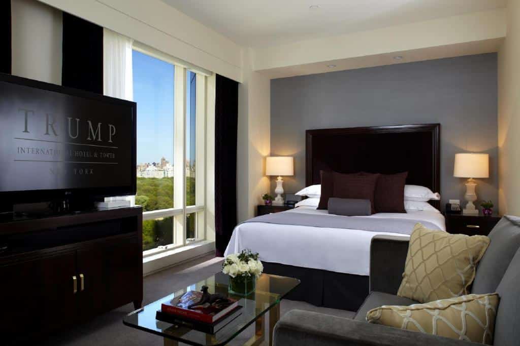 No quarto do Trump International New York há uma cama de casal ao centro, com cabeceira escura e almofadas decorativas. À frente, um sofá cinzento e uma mesa de centro de vidro; à esquerda, uma grande TV. Ao fundo, uma janela ampla com vista para a cidade.