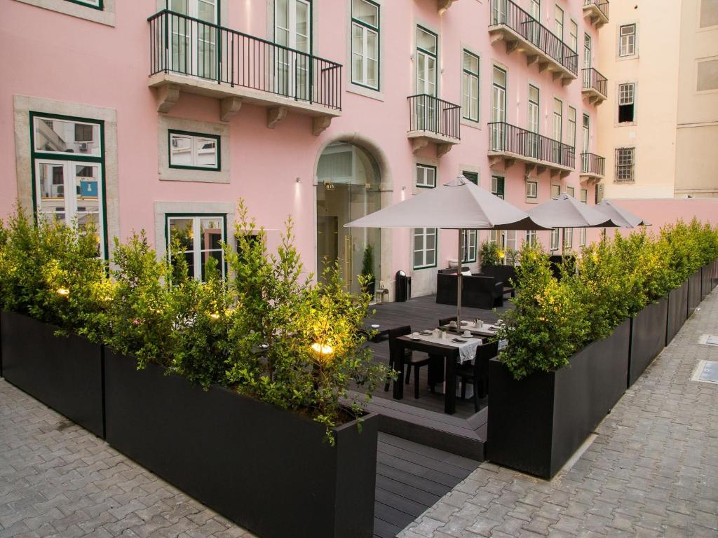 Parte da fachada do Portugal Boutique Hotel, com paredes cor-de-rosa claro, varandas de ferro e janelas amplas, além de mesas com guarda-sóis junto à entrada.