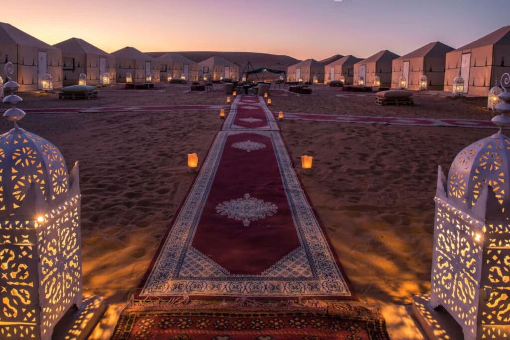 Acampamento elegante no deserto de Merzouga ao anoitecer, com lanternas ornamentadas, um caminho alcatifado e tendas iluminadas ao fundo.