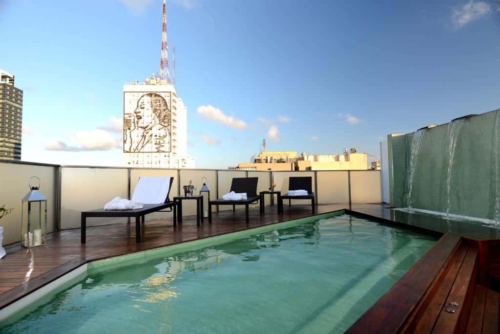 piscina no terraço do Eurobuilding Hotel Boutique Buenos Aires, retangular, com borda de madeira e parede de vidro, espreguiçadeiras escuras com toalhas brancas e vista panorâmica da cidade