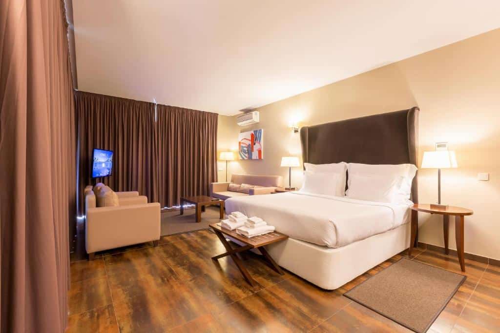 Quarto do NAU Salgados Palm Village – All Inclusive com cama de casal do lado direito da imagem no centro do quarto com uma cômoda em cada lado da cama com luminária, no pé da cama uma cômoda de madeira com toalhas. Do lado esquerdo da imagem um sala de estar com sofá. Representa hotéis all inclusive em Portugal.