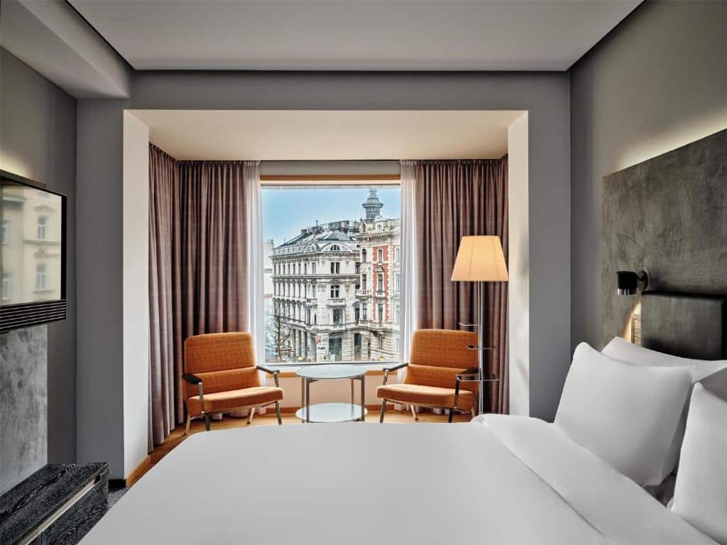 Quarto no Radisson Blu Das Triest Hotel, com cama grande, duas poltronas junto a uma janela, um candeeiro de pé e vista para um edifício da cidade.