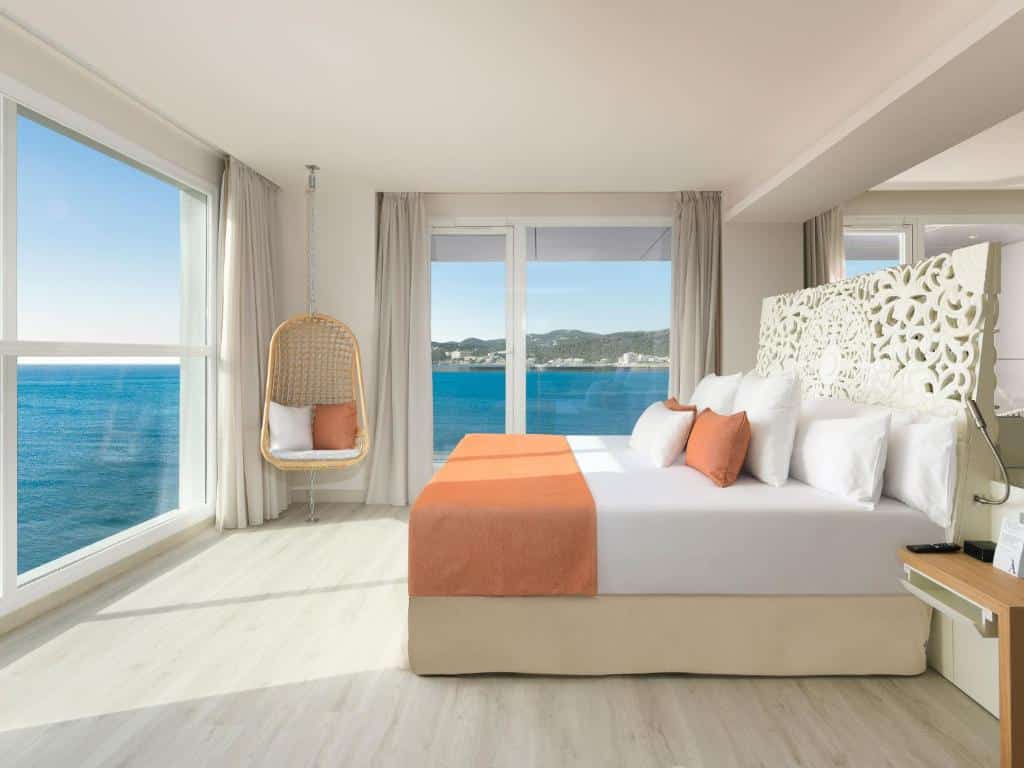 Quarto moderno e bem iluminado do Amàre Beach Hotel Ibiza Adults Only, com janelas grandes, vista para o mar, uma cama com detalhes em laranja e uma cadeira suspensa.