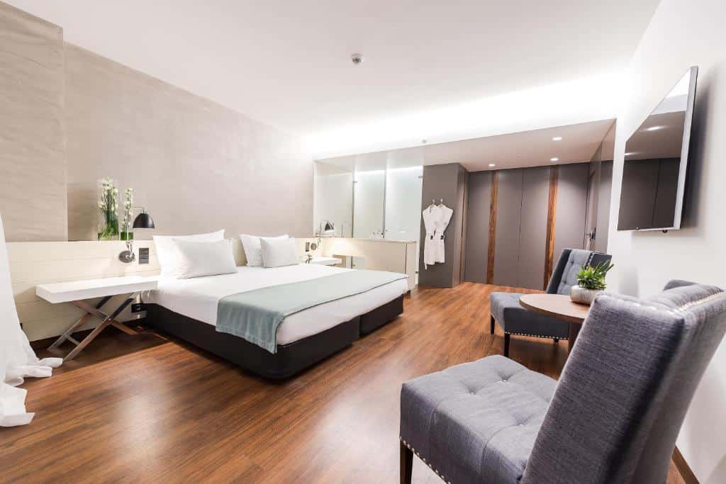 Quarto moderno no Lux Lisboa Park com uma cama de casal, duas cadeiras, uma mesa pequena, TV na parede e uma área de roupeiro aberta.