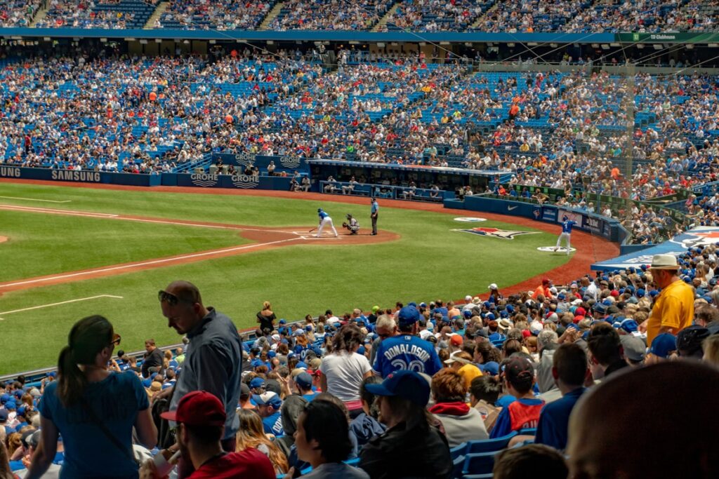 Estádio cheio de adeptos a assistir a uma partida de basebol, com um jogador a lançar a bola e o árbitro atrás da base