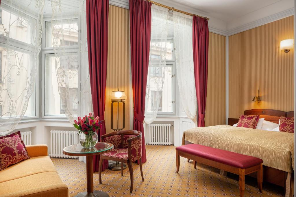 Quarto do Hotel Paris Prague, com cama de casal, cortinas vermelhas, um sofá, uma mesa com cadeiras e janelas grandes com cortinados transparentes.