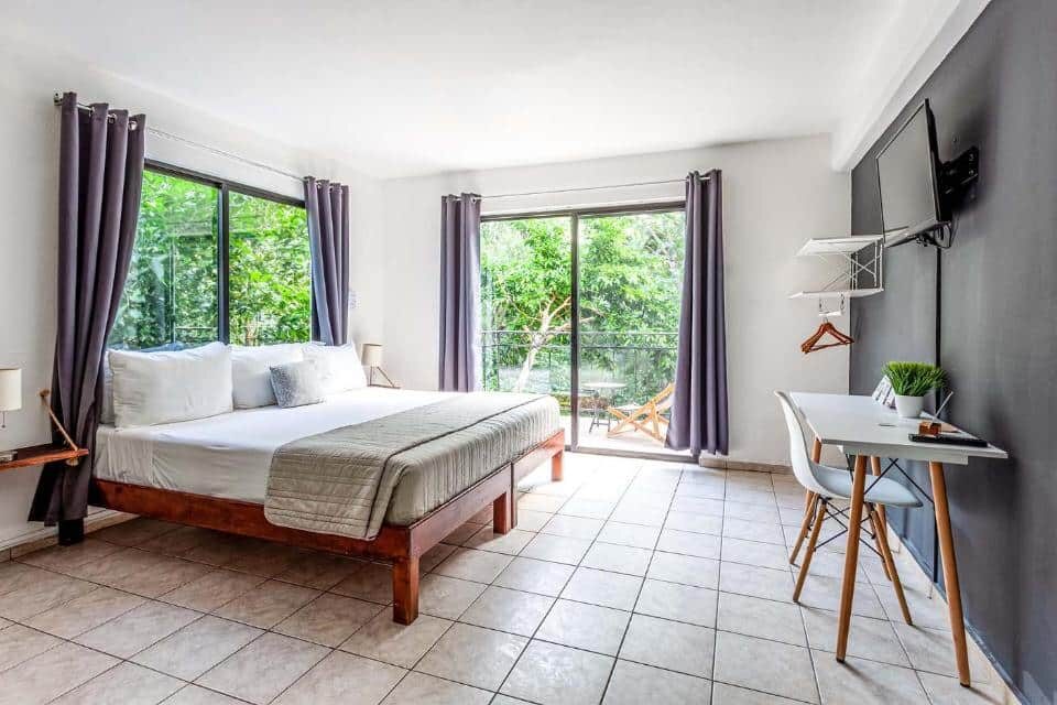 Quarto do Che Suites Playa com cama de casal ao centro, secretária e vista para área verde pelas portas de vidro da varanda.