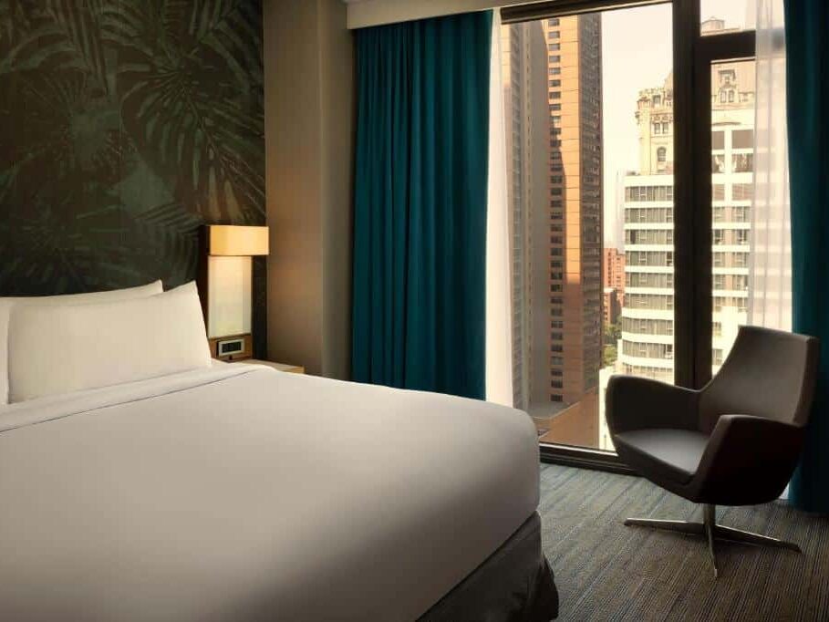 No quarto do The Cloud One New York Downtown há uma cama de casal com lençóis brancos ao centro. À direita, uma cadeira de leitura preta junto à janela; à esquerda, uma mesa de cabeceira com candeeiro e despertador. Ao fundo, uma janela grande com cortinados azuis.