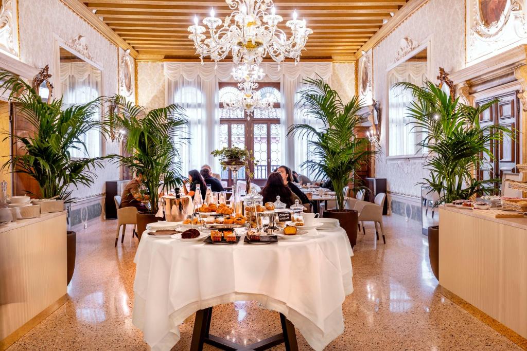 Sala de jantar luxuosa num edifício histórico, com móveis de madeira, arabescos, lustres de cristal e plantas ornamentais, e grupos de pessoas a desfrutar de um banquete