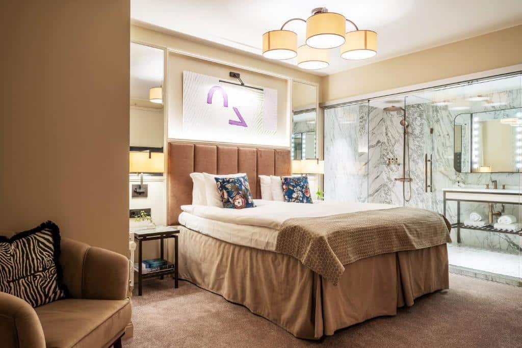 Quarto do Bank Hotel com poltrona, cama de casal e casa de banho com portas de vidro.