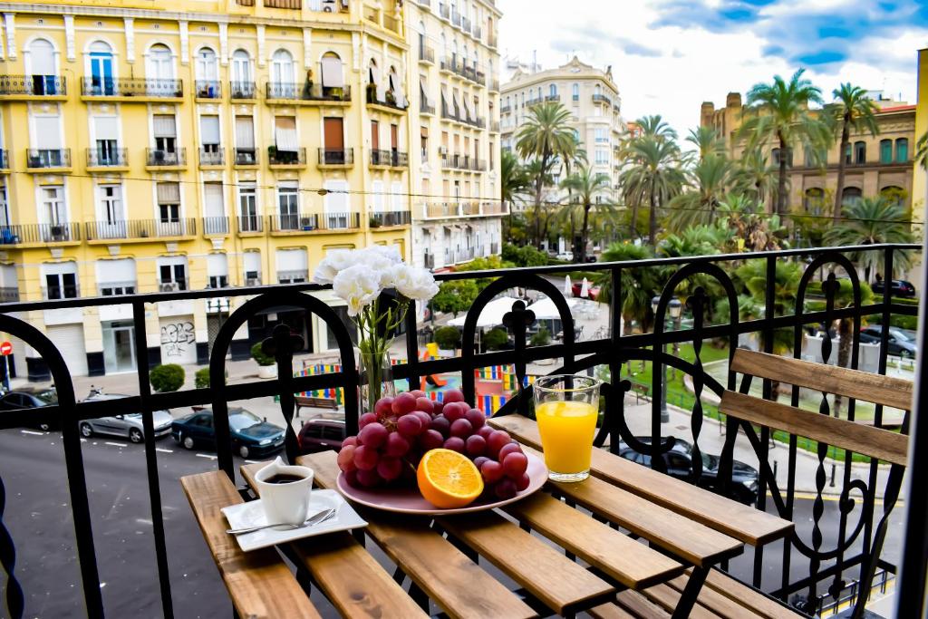 Varanda do Valencia Apartamentos Goya com vista para edifícios amarelos e palmeiras, com mesa de madeira, fruta fresca, sumo de laranja e café numa chávena sobre o balcão.