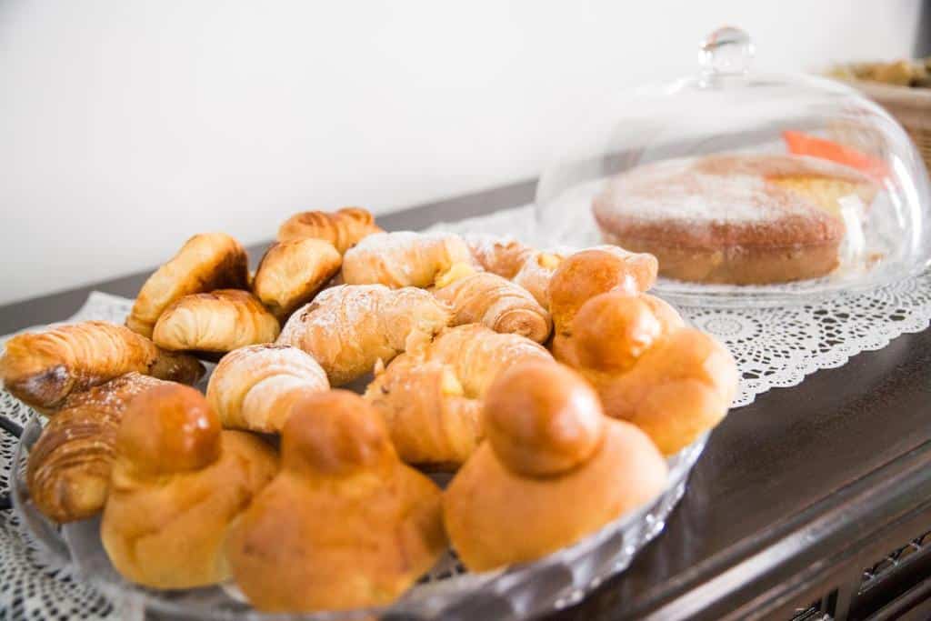 Variedade de pães e doces dispostos numa mesa com toalha rendada, incluindo croissants, argolas, bolos e outros itens, no NapoliMia Boutique Hotel.