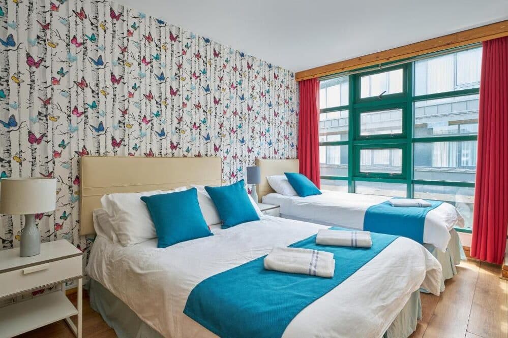 Quarto do Abigail's Hostel com paredes decoradas com borboletas coloridas, duas camas de casal com almofadas e cobertores azuis, candeeiro dourado e cortinas vermelhas na janela com vista para a cidade.