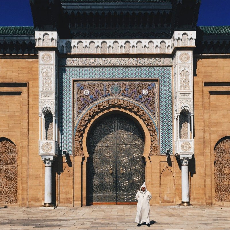 Uma pessoa em trajes tradicionais caminha em frente a uma grande porta ornamentada, com azulejos intrincados e pilares decorativos, em Casablanca.