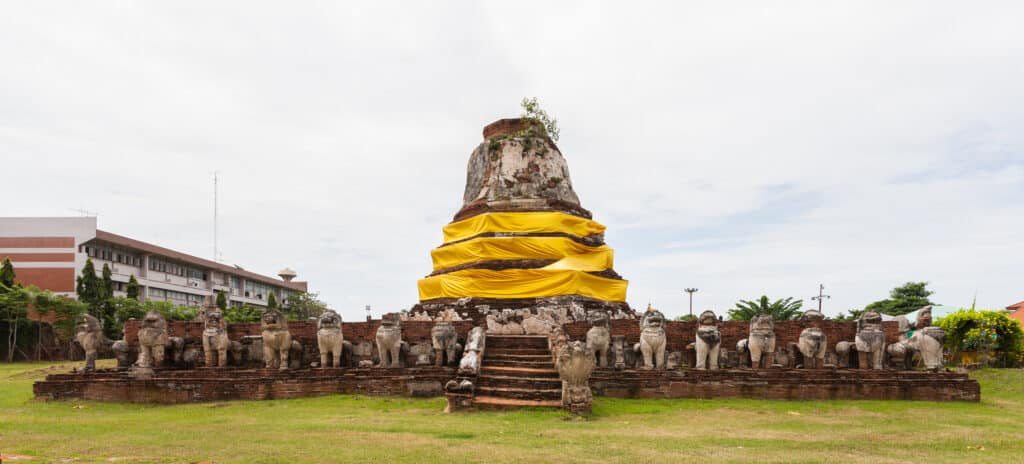 Antigo pagode de pedra envolto em tecido amarelo, rodeado por estátuas de leões, num relvado, em Ayutthaya.