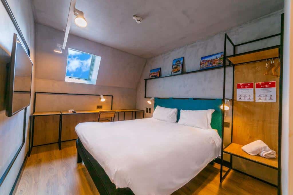 Quarto moderno do ibis Porto Centro Mercado Bolhão com cama de casal, chão de madeira, secretária, arte na parede e uma janela com céu azul.