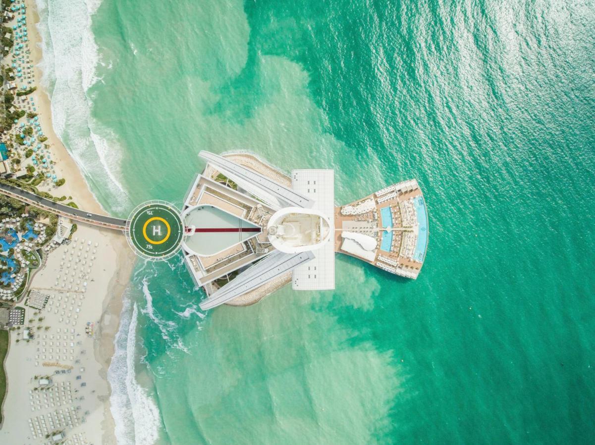 Vista aérea do Burj Al Arab em forma de vela sobre o mar, rodeado por águas azul-turquesa, com heliporto e zona de praia.