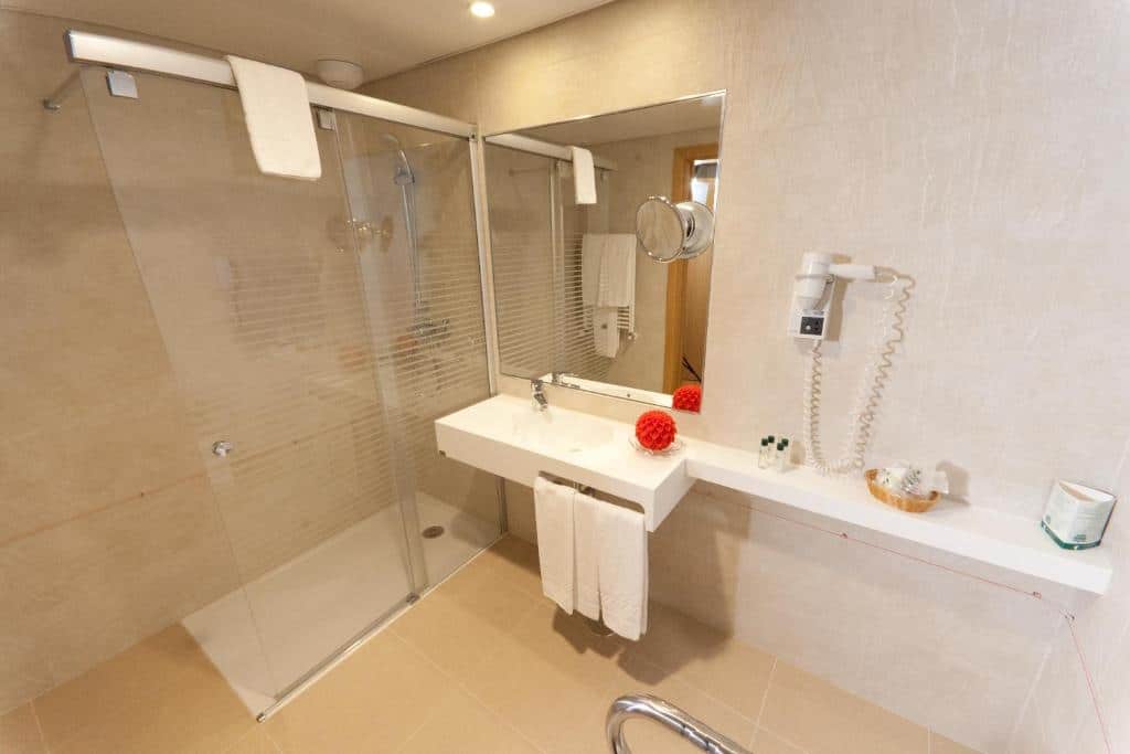 Casa de banho moderna com chuveiro de vidro, lavatório branco, toalhas, secador de cabelo na parede e produtos de higiene no balcão, no Hotel Cinquentenário & Conference Center.