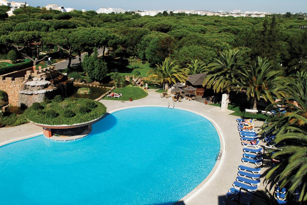 Ampla piscina exterior rodeada de espreguiçadeiras, palmeiras e vegetação, com edifícios visíveis ao longe, no Falésia Hotel – Adults Only em Albufeira.
