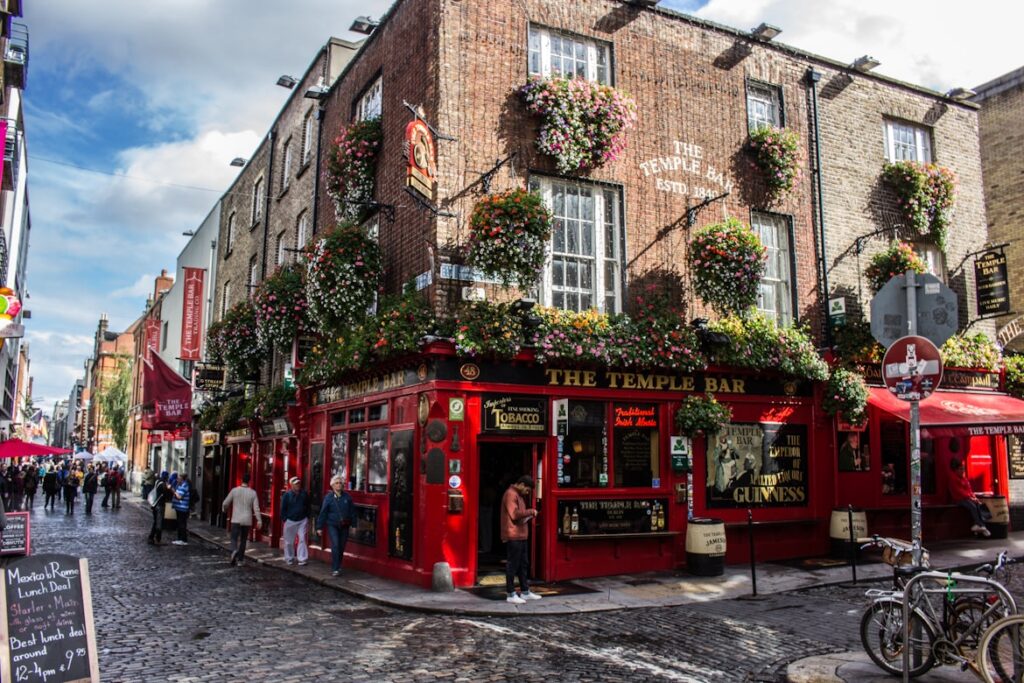 Rua no bairro de Temple Bar, em Dublin, com fachadas de tijolo vermelho decoradas com floreiras coloridas. Na esquina, o famoso The Temple Bar, com a sua decoração tradicional em vermelho, e pessoas a passar na rua movimentada.