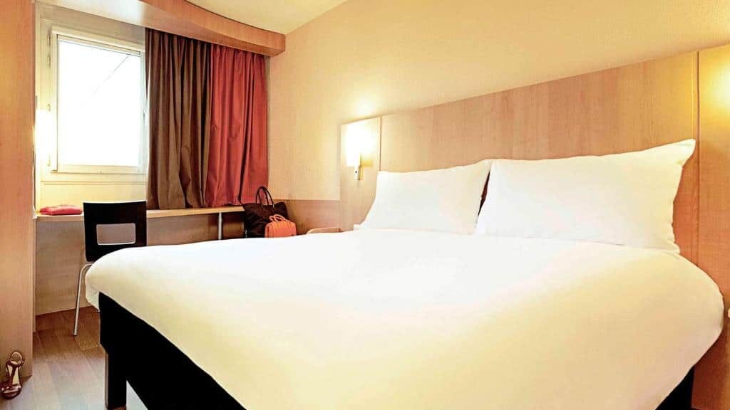 Quarto do Hotel Ibis Lisboa José Malhoa com cama de casal, cabeceira de madeira, candeeiro de parede e secretária com cadeira.