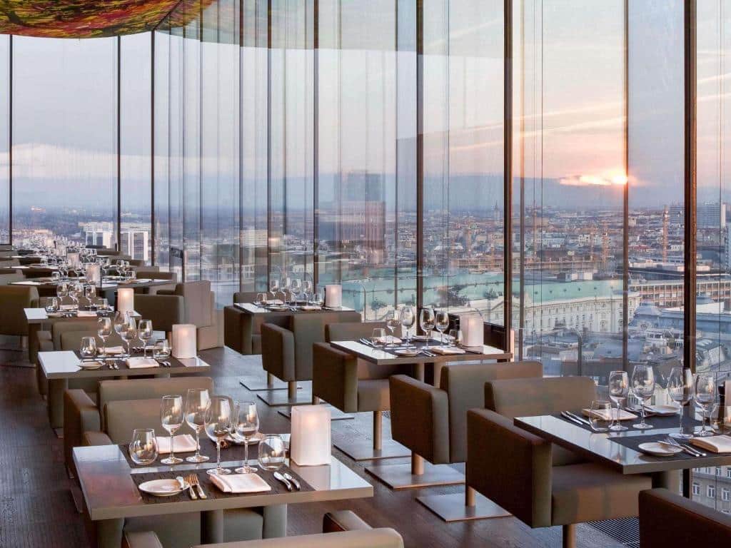 Restaurante moderno no SO/ Vienna, com janelas do chão ao teto, mesas postas e vista da cidade ao pôr do sol.