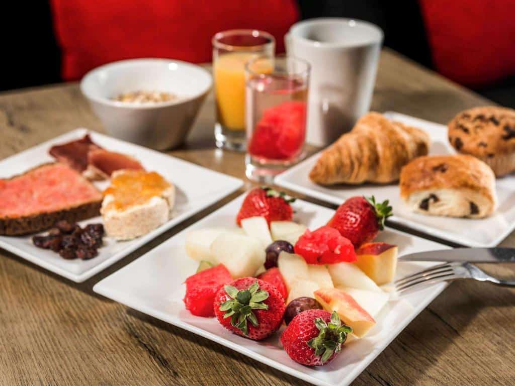 Pratos de pequeno-almoço do Hotel Ibis Lisboa Saldanha com fruta variada, doces, pão e sumos sobre uma mesa de madeira.