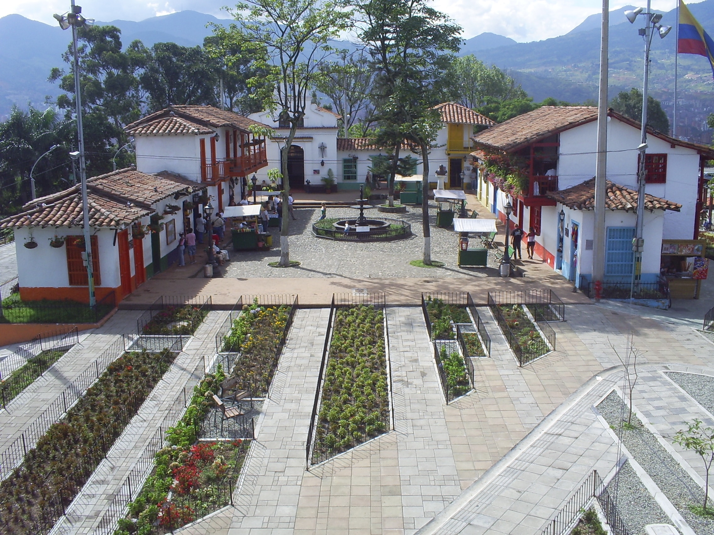 Vista aérea do Pueblito Paisa, com jardins, edifícios coloridos, uma fonte central e montanhas ao fundo.