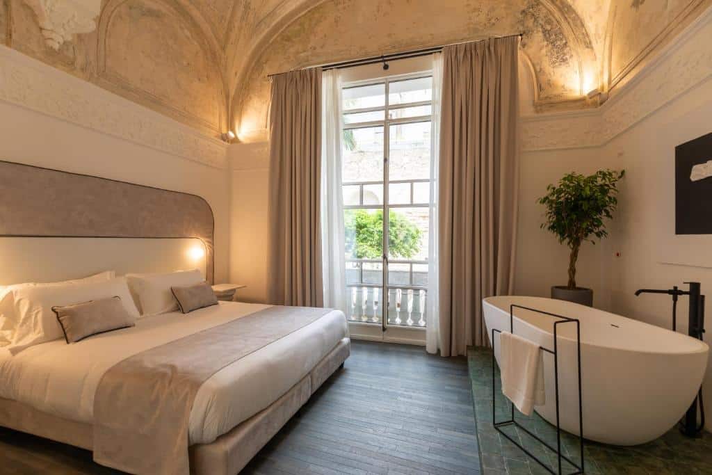 Quarto luxuoso do Artemisia Domus com paredes de tijolo à vista, cama king size com cabeceira estofada, mesa de centro de vidro e banheira redonda junto à janela com vista para o jardim.