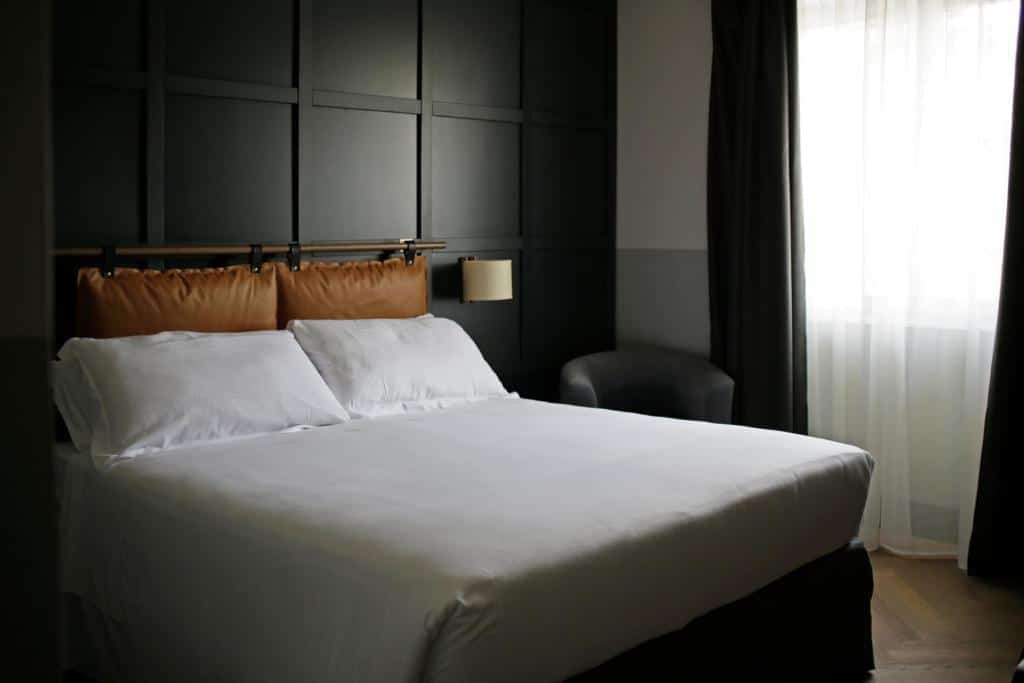quarto do c-hotels Club House Roma, um dos hotéis baratos em Roma, com cama de casal grande, mesa de cabeceira com candeeiro de ambos os lados, parede com textura, um cadeirão ao lado e uma janela grande com cortinados escuros