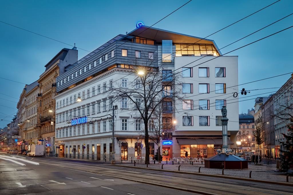 Fachada do Radisson Blu Das Triest Hotel, de vários andares, com grandes janelas e um telhado de vidro, numa rua da cidade à noite.