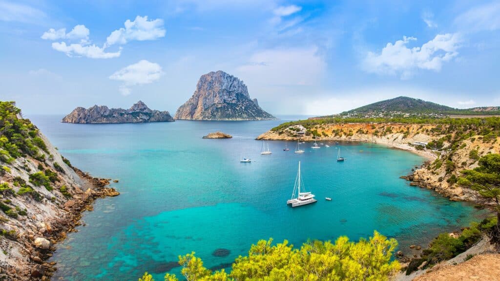 Baía azul-turquesa em Ibiza com veleiros ancorados, costa rochosa, colinas verdes e uma grande ilha ao fundo sob um céu azul.