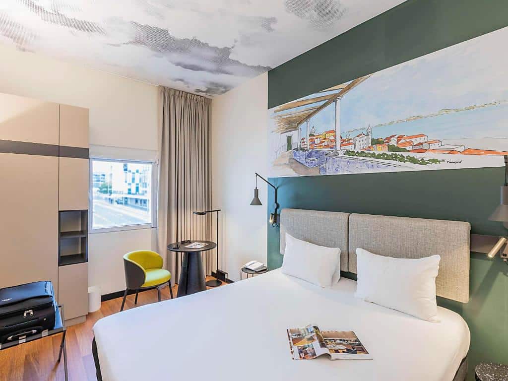 Quarto do Ibis Lisboa Parque das Nações com uma janela com cortinas, um pequeno armário, uma cama de casal, uma mesinha com uma poltrona e candeeiros nas laterais da cama